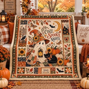 Peut inclure: Une courtepointe rectangulaire représentant un fantôme central portant un chapeau de sorcière en patchwork et tenant une lanterne, avec un chat noir, des citrouilles, des tournesols et des feuilles d'automne. Le texte indique Cozy Spooky Nights, Trick or Treats et BOO.
