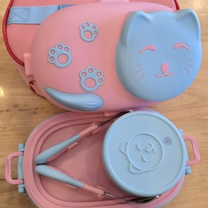 May include: Pink and blue cat-shaped lunch box set. The set includes a pink insulated bag, a pink lunch box with a blue cat face, and a matching tray with utensils and a small container. The lunch box has paw print details.
