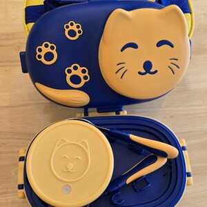 May include: A blue and yellow cat-themed lunch box set. The set includes a lunch box with a cat face design, a matching lid, and utensils. The lunch box has a yellow handle and a matching yellow carrying case.