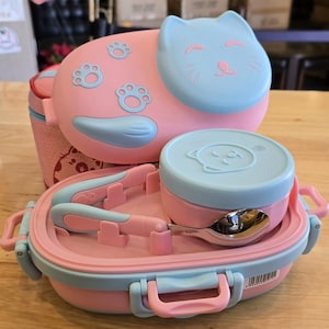 May include: A pink and blue cat-shaped lunch box set. The set includes a cat-shaped container, a round container, a spoon, and a pair of tongs. The lunch box has a pink base and a blue lid. The set is designed for children.