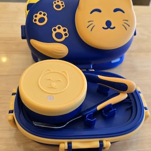 May include: A blue and yellow cat-themed lunch box set. The set includes a lunch box, a round container, a fork, and a spoon. The lunch box has a cat face design on the lid and paw prints on the body.