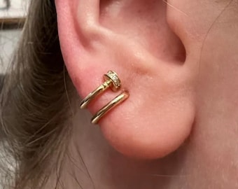 Puede incluir: Un pendiente tipo ear cuff en tono dorado con un diseño de doble banda que rodea el cartílago. La banda superior presenta un pequeño detalle circular incrustado con piedras transparentes que recuerda a una cabeza de clavo, y la banda inferior es lisa.