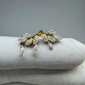 May include: A gold-toned V-shaped ear cuff or earring featuring marquise-cut clear stones arranged in a leaf-like pattern. Several marquise-cut stones dangle from the main structure, creating a cascading effect with a bright, reflective finish.