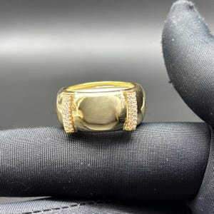 Könnte beinhalten: Ein goldfarbener Ring mit einem breiten Band und zwei vertikalen Abschnitten aus kleinen, klaren Edelsteinen. Der Ring hat eine polierte, reflektierende Oberfläche und eine abgerundete Form. Der Ring wird auf einer schwarzen Oberfläche präsentiert.