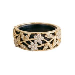 Op de afbeelding: Een goudkleurige ring met een bloemmotief. De ring heeft een zwart interieur en is versierd met diamantachtige stenen in een bloemen- en bladerenpatroon. De band is ongeveer 1,3 cm breed.