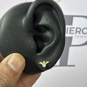 Könnte beinhalten: Ein goldfarbener Ohrstecker in Fledermausform mit einem klaren, runden Edelstein in der Mitte. Der Ohrring wird auf einem schwarzen Ohrmodell gezeigt. Die Flügel der Fledermaus sind ausgebreitet.