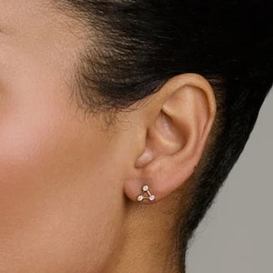 May include: A gold stud earring with three small, round, clear gemstones arranged in a triangular shape. The earring is set against the skin of a person's ear.