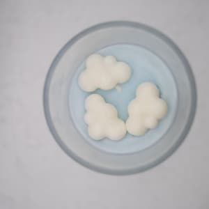 May include: A clear glass bowl with a light blue interior holds three white cloud-shaped wax melts. The bowl sits on a white surface, creating a clean and minimalist aesthetic. The melts are clustered together.