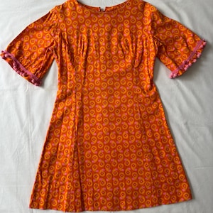 May include: An orange dress featuring a repeating pattern of pink and yellow circles. The dress has short sleeves trimmed with pink pom-poms and a fitted bodice that flares at the waist. The fabric is lightweight.