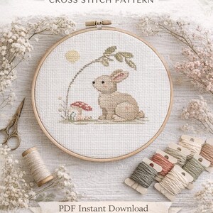 May include: A cross-stitch pattern featuring a beige rabbit, mushroom, and sun design inside a wooden embroidery hoop. The text "Spring Rabbit" and "Cross Stitch Pattern" are at the top. The words "PDF Instant Download" and "Pure Loop Pattern" are also visible.