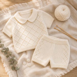 May include: A cream-colored short-sleeved knit shirt with a rounded collar and a diamond-patterned texture, paired with matching knit shorts featuring side diamond detailing. The set is accompanied by a ball of yarn and two knitting needles.