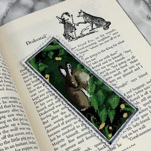 May include: A handmade bookmark featuring a cross-stitch design of a brown hare in a green foliage setting. The bookmark has a white lace trim and is placed on an open book. The text "Drakestail" is visible.