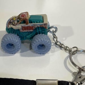 K&ouml;nnte beinhalten: Ein Miniatur-Monster-Truck-Spielzeug mit dem Wort "ZOMBIE" an der Seite. Der Truck hat blaue R&auml;der, einen t&uuml;rkisfarbenen K&ouml;rper und ein beiges Oberteil. Eine silberne Kette und ein Schl&uuml;sselring sind am Truck befestigt.
