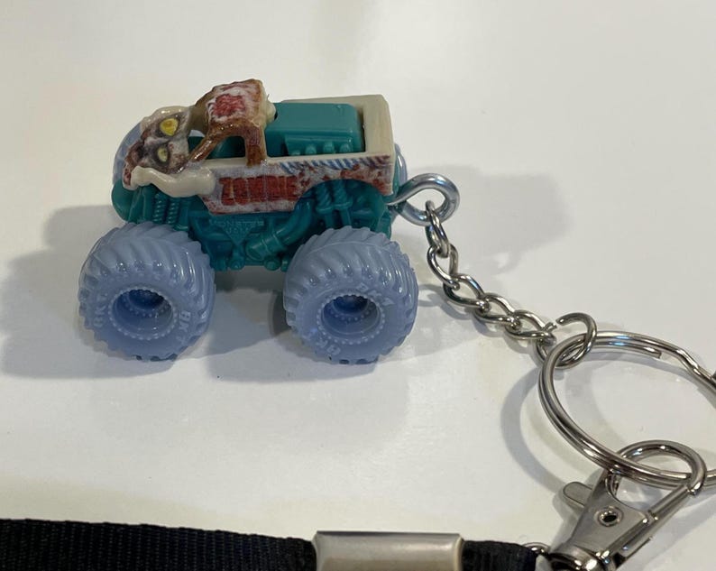 K&ouml;nnte beinhalten: Ein Miniatur-Monster-Truck-Spielzeug mit dem Wort "ZOMBIE" an der Seite. Der Truck hat blaue R&auml;der, einen t&uuml;rkisfarbenen K&ouml;rper und ein beiges Oberteil. Eine silberne Kette und ein Schl&uuml;sselring sind am Truck befestigt.