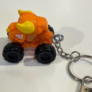 K&ouml;nnte beinhalten: Orangener und gelber Monster-Truck-Spielzeug-Schl&uuml;sselanh&auml;nger mit schwarzen R&auml;dern. Der Truck hat ein bullenartiges Design mit dem Text "El Toro Loco". Der Schl&uuml;sselanh&auml;nger enth&auml;lt eine silberne Kette und einen Schl&uuml;sselring.