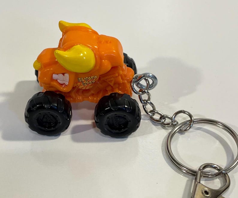 K&ouml;nnte beinhalten: Orangener und gelber Monster-Truck-Spielzeug-Schl&uuml;sselanh&auml;nger mit schwarzen R&auml;dern. Der Truck hat ein bullenartiges Design mit dem Text "El Toro Loco". Der Schl&uuml;sselanh&auml;nger enth&auml;lt eine silberne Kette und einen Schl&uuml;sselring.