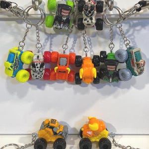 K&ouml;nnte beinhalten: Eine Sammlung von Spielzeug-Monster-Truck-Schl&uuml;sselanh&auml;ngern in verschiedenen Farben, darunter Gelb, Orange, Gr&uuml;n und Blau. Jeder Schl&uuml;sselanh&auml;nger zeigt ein Miniatur-Monster-Truck-Design mit schwarzen R&auml;dern und ist an einer silbernen Kette befestigt. Einige Trucks haben "Grave Digger"-Grafiken.
