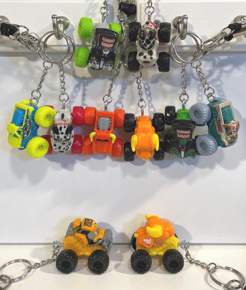 K&ouml;nnte beinhalten: Eine Sammlung von Spielzeug-Monster-Truck-Schl&uuml;sselanh&auml;ngern in verschiedenen Farben, darunter Gelb, Orange, Gr&uuml;n und Blau. Jeder Schl&uuml;sselanh&auml;nger zeigt ein Miniatur-Monster-Truck-Design mit schwarzen R&auml;dern und ist an einer silbernen Kette befestigt. Einige Trucks haben "Grave Digger"-Grafiken.