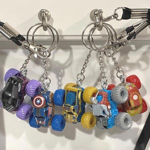 May include: Six toy monster truck keychains in various colors, including purple, blue, yellow, red, and silver. Each keychain features a different truck design, with details like the Captain America shield and Iron Man's armor.