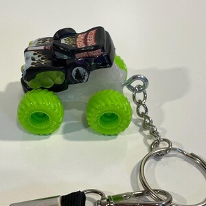 K&ouml;nnte beinhalten: Ein Miniatur-Grave Digger Monster Truck Schl&uuml;sselanh&auml;nger. Der Truck ist schwarz mit gr&uuml;nen und wei&szlig;en Akzenten, mit Flammen und dem Grave Digger Logo. Die Reifen sind hellgr&uuml;n. Der Schl&uuml;sselanh&auml;nger hat eine silberne Kette und einen Ring.