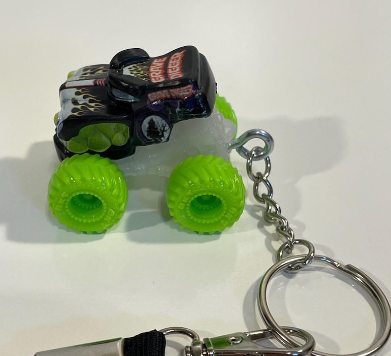 K&ouml;nnte beinhalten: Ein Miniatur-Grave Digger Monster Truck Schl&uuml;sselanh&auml;nger. Der Truck ist schwarz mit gr&uuml;nen und wei&szlig;en Akzenten, mit Flammen und dem Grave Digger Logo. Die Reifen sind hellgr&uuml;n. Der Schl&uuml;sselanh&auml;nger hat eine silberne Kette und einen Ring.
