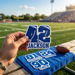 May include: A blue and white embroidered patch featuring the number 42 and the name JACKSON, with additional patches labeled 81 RODRIGUEZ, 24 NGUYEN, and 90 CHEN displayed on a blue towel alongside a brown leather football on a wooden bench.