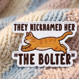 May include: An embroidered patch with a white background featuring a running orange tabby cat. The text above the cat reads "THEY NICKNAMED HER" and the text below the cat reads "THE BOLTER" in dark brown capital letters with a scalloped border.