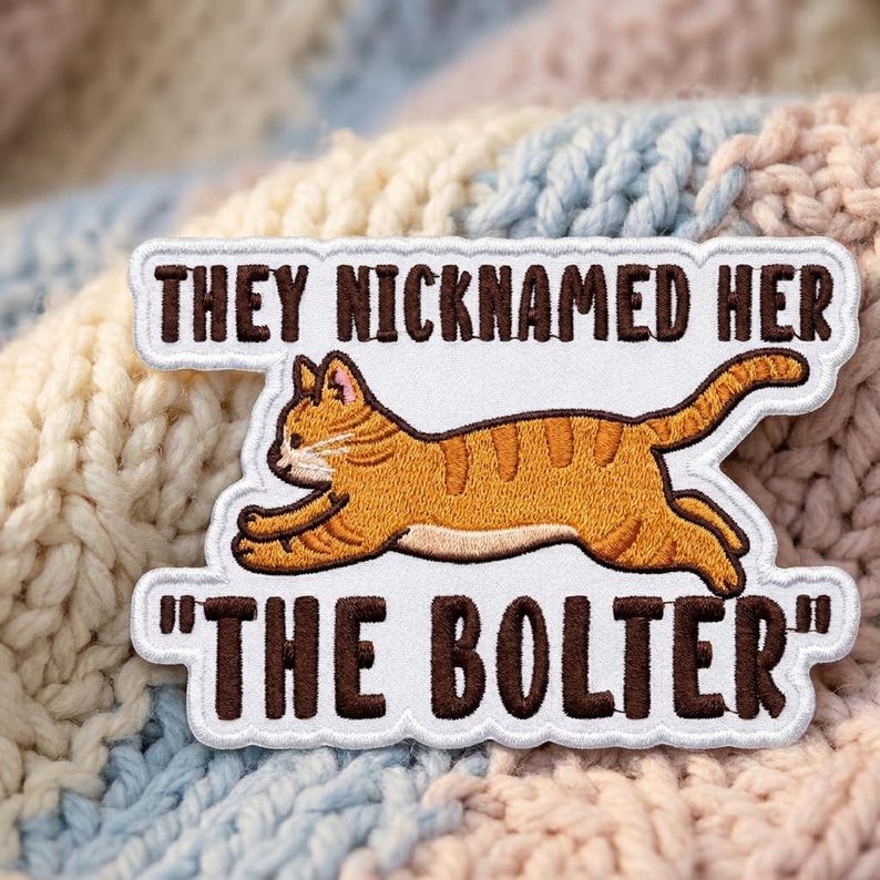 May include: An embroidered patch with a white background featuring a running orange tabby cat. The text above the cat reads "THEY NICKNAMED HER" and the text below the cat reads "THE BOLTER" in dark brown capital letters with a scalloped border.