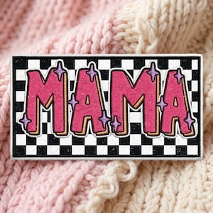 May include: A rectangular embroidered patch featuring a black and white checkered background. The word MAMA is written in large, bold pink letters with a tan outline. Small, light purple four-pointed star shapes are scattered around the letters.