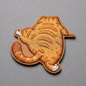 May include: An embroidered patch shaped like a ginger tabby cat viewed from behind. The cat has orange fur with darker orange stripes, a curled tail, and small white markings on its paws. The patch features detailed stitching throughout.