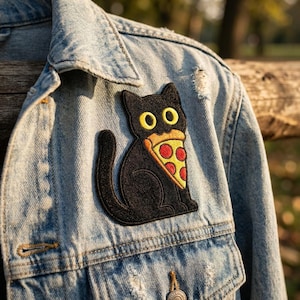 May include: A light blue denim jacket featuring an embroidered patch of a black cat with wide yellow eyes holding a slice of pepperoni pizza in its mouth. The jacket has a classic collar, metal buttons, and visible stitching along the seams.