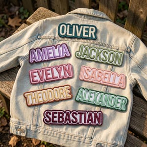 May include: A light-wash denim jacket featuring seven embroidered name patches in various colors. The patches display the names OLIVER, AMELIA, JACKSON, EVELYN, ISABELLA, THEODORE, ALEXANDER, and SEBASTIAN in bold, capitalized, sans-serif lettering.