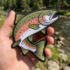 May include: An embroidered iron-on patch shaped like a rainbow trout. The fish features an olive green back with black spots, a white underbelly, and a distinct pinkish-red stripe running along its side. It has a black border and fins.