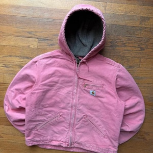 May include: A pink hooded jacket with a full zipper closure and multiple pockets. The jacket has a gray fleece-lined hood and a small logo patch on the chest. The jacket is made of a durable, workwear-style fabric.