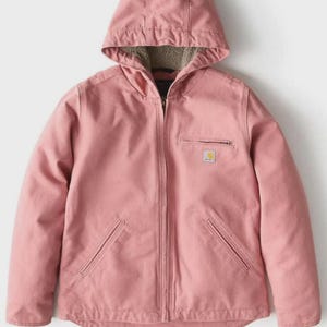 May include: A dusty rose hooded jacket with a full-length zipper and two front pockets. The jacket has a small square logo on the chest and a soft, fuzzy lining visible inside the hood. The jacket is made of a durable, workwear-style material.