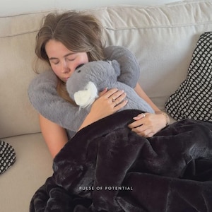 May include: A grey plush elephant neck pillow and a black faux fur blanket. The pillow is being held by a person. The blanket is draped over the person. The text "PULSE OF POTENTIAL" is visible.