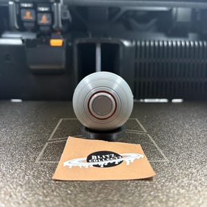 May include: A gray, spherical object with a red ring detail, resting on a black base. A small, tan card with a black and white logo is in front of the sphere. The logo reads "BLITZ COLLECT".