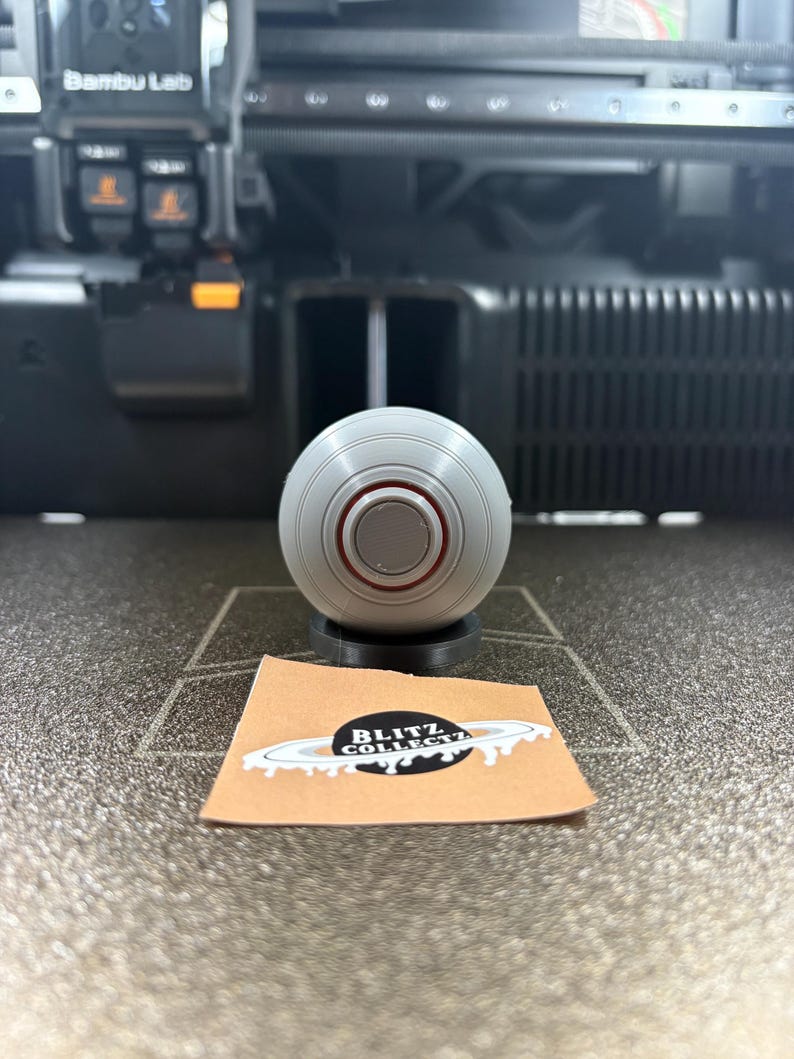 May include: A gray, spherical object with a red ring detail, resting on a black base. A small, tan card with a black and white logo is in front of the sphere. The logo reads "BLITZ COLLECT".