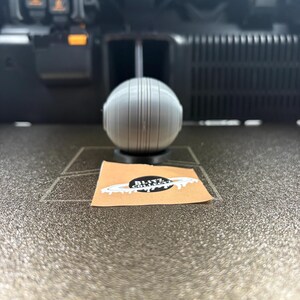 May include: A gray, spherical object with vertical lines, possibly a 3D-printed item, sits on a dark surface. A small sticker with the text "Blitz Collect" and a planet design is visible. The background shows a 3D printer.