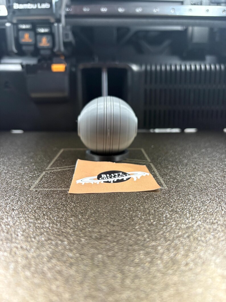 May include: A gray, spherical object with vertical lines, possibly a 3D-printed item, sits on a dark surface. A small sticker with the text "Blitz Collect" and a planet design is visible. The background shows a 3D printer.