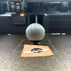 May include: A gray, spherical 3D-printed object with vertical lines, resting on a black base. A small, tan sticker with the text "BLITZ COLLECT" is in front of the object. The background includes a 3D printer.
