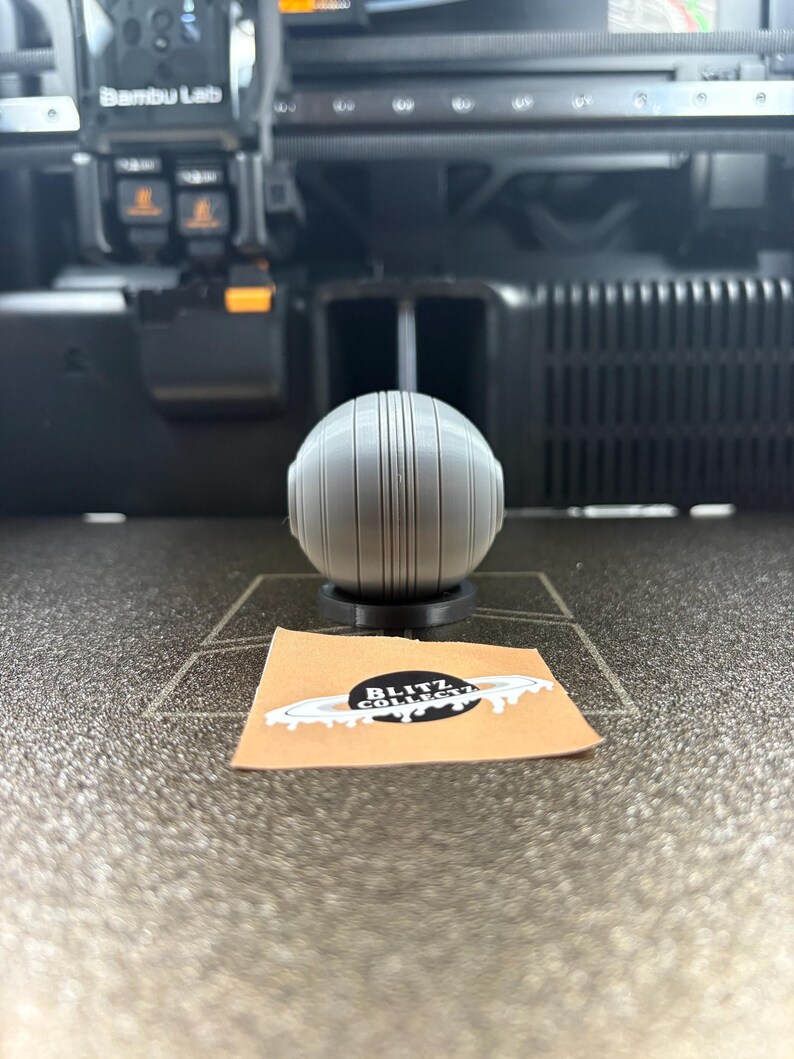 May include: A gray, spherical 3D-printed object with vertical lines, resting on a black base. A small, tan sticker with the text "BLITZ COLLECT" is in front of the object. The background includes a 3D printer.