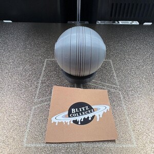 May include: A gray, spherical object with vertical lines and a small base. A brown square card with the text "BLITZ COLLECTZ" and a planet design is in front of the sphere.