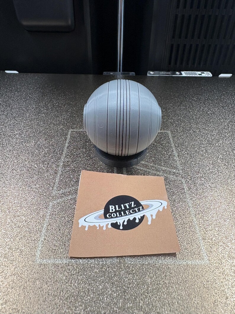 May include: A gray, spherical object with vertical lines and a small base. A brown square card with the text "BLITZ COLLECTZ" and a planet design is in front of the sphere.