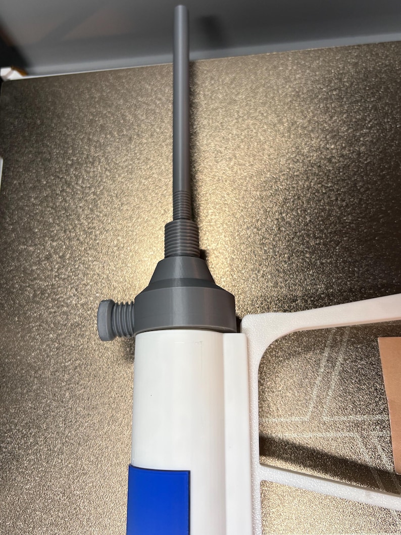 Op de afbeelding: Een witte en blauwe fles met een grijze tuit en een lange, grijze, cilindrische verlenging. De fles heeft een blauw rechthoekig etiket. De tuit heeft een schroefdraadbasis en een kleine, cilindrische knop aan de zijkant.