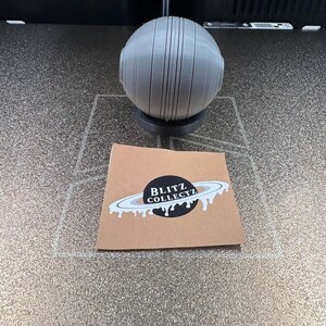 May include: A gray, spherical object with vertical lines and two circular protrusions, resting on a black base. A small card with the text "BLITZ COLLECTZ" and a planet design is in the foreground.