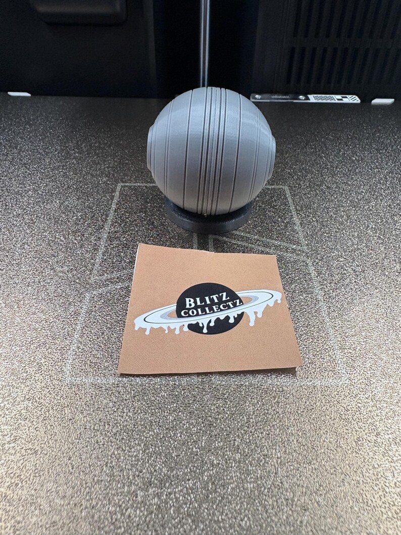 May include: A gray, spherical object with vertical lines and two circular protrusions, resting on a black base. A small card with the text "BLITZ COLLECTZ" and a planet design is in the foreground.