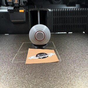 May include: A gray and red spherical object sits on a black base, positioned in front of a 3D printer. A small brown card with the text "BLITZ COLLECT" is also visible.