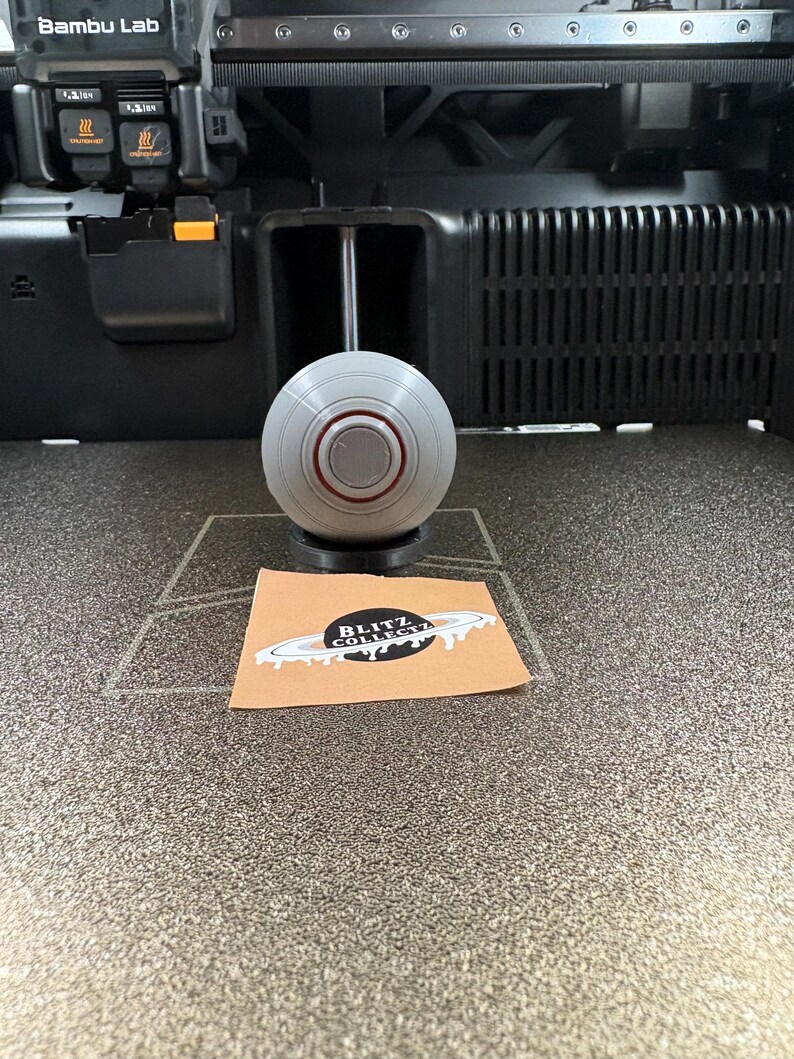 May include: A gray and red spherical object sits on a black base, positioned in front of a 3D printer. A small brown card with the text "BLITZ COLLECT" is also visible.