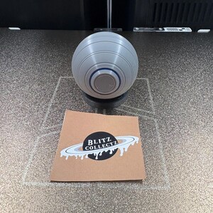May include: A gray, spherical object with a circular base, featuring concentric rings and a central button. A small, brown card with the text "BLITZ COLLECTZ" and a planet graphic is also present.